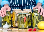 Forskellige glas med hjemmelavede syltede agurker, hvidløg, chili og krydderier. Glas med metal- og papirlåg, præsenteret på et træbord med friske ingredienser.