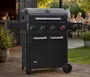 Sort gasgrill med fire kontrolknapper, to sidehylder, dobbeltlåge opbevaringsskab og termometer på låget. Robuste hjul for nem flytning. Moderne, stilrent udendørs design.
