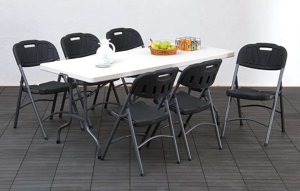 Rektangulært hvidt klapbord med seks sorte klapstole. Bordet har en frugtkurv, drikkevarer og et tekandesæt. Stolene har metalstel og formede plastiksæder.