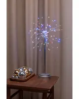 Bordslampa formad som fyrverkeri med flerfärgade LED-lampor på tunna trådar, silverfot och stång. Dekorativa discobollar i guld och silver på träbricka bredvid.