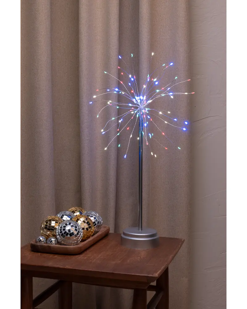 Bordslampa formad som fyrverkeri med flerfärgade LED-lampor på tunna trådar, silverfot och stång. Dekorativa discobollar i guld och silver på träbricka bredvid.