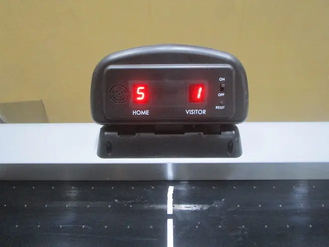 Digitalt airhockey-scoreboard med rød LED-skærm, viser hjem- og gæstescorer. Har tænd/sluk-knap, reset-knap og monteres på bordet. Kompakt sort plastdesign.