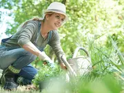 Kvinde med solhat, handsker og gummistøvler knæler på en havepude og planter udendørs. Inkluderer vandkande og potteplante, ideel til havearbejde.