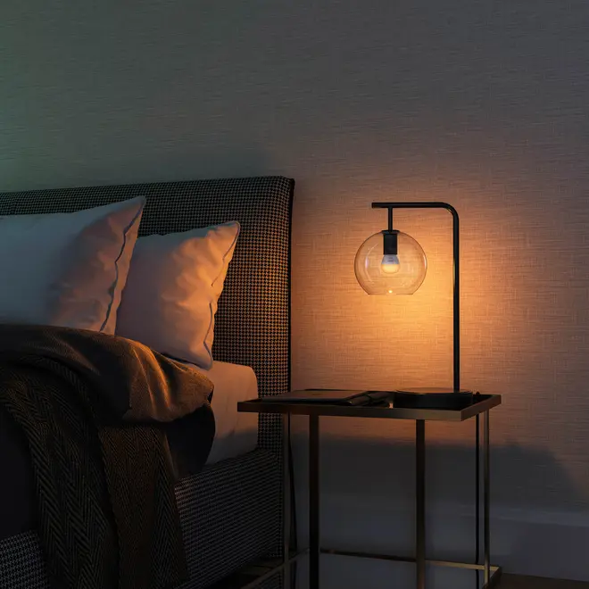Moderne natbordslampe med sort metalstel og klar glaskuppel, placeret på et firkantet sidebord. Udsender varmt lys, ideel til soveværelser eller stuer.