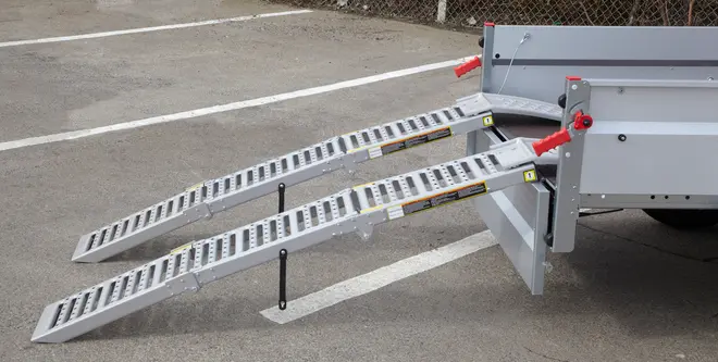 Sæt kraftige metalramper monteret på trailer, med perforeret overflade for greb, støtteben for stabilitet og røde håndtag til fastgørelse. Velegnet til lastning af køretøjer eller udstyr.