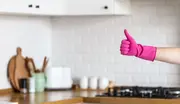 En person med en starkt rosa gummihandske för städning ger tummen upp i ett modernt kök med vita kakel och träbänkskivor.