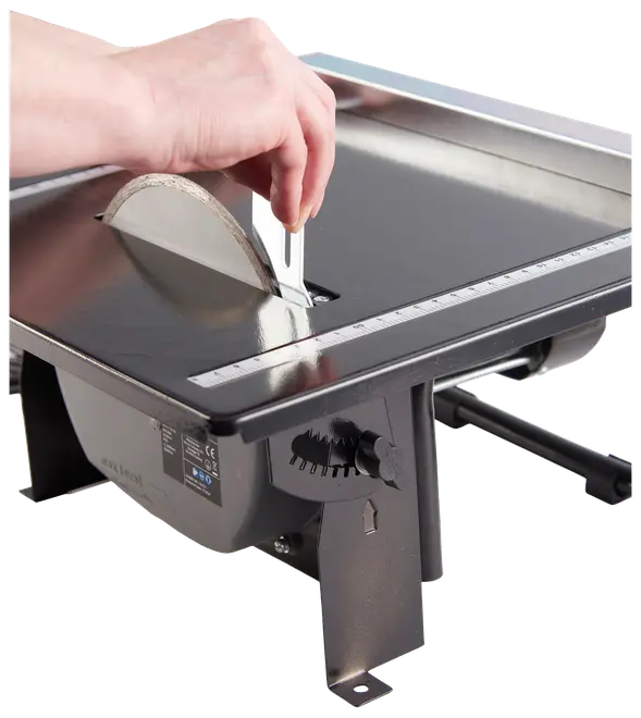 Elektrisk fliseskærer med rund klinge, justerbar skærestyr og måleskala på flad metaloverflade. Hånd justerer styret. Velegnet til præcis fliseskæring.