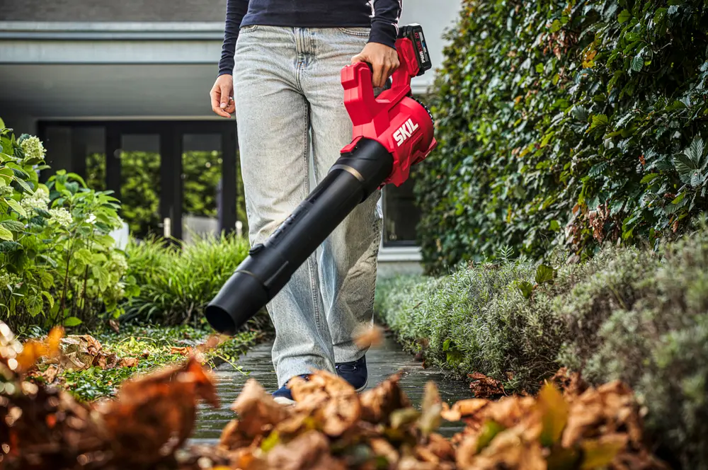 Röd SKIL batteridriven lövblås med ergonomiskt handtag, lätt design och lång svart munstycke, visas i bruk för att blåsa bort löv från en trädgårdsgång.