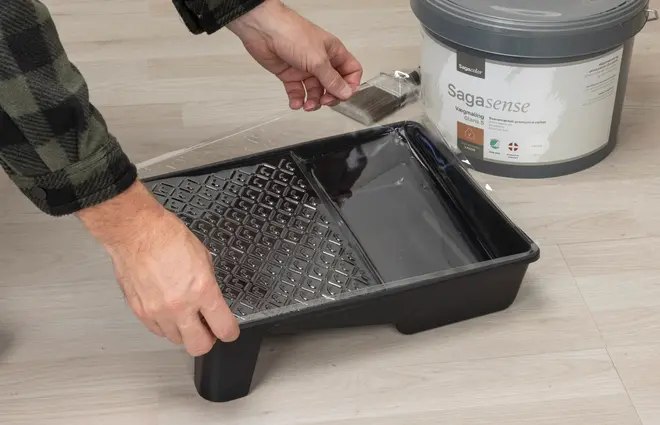 Sort plastik malerbakke med struktureret rulleflade og glat malingsrum, vist sammen med malerspand og pensel.