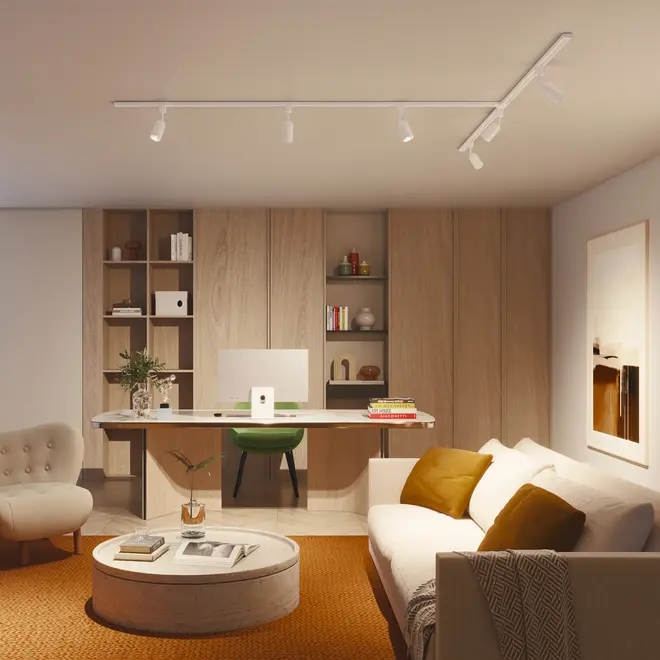 Moderne hjemmekontor med hvidt skrivebord, grøn stol, beige sofa med brune puder, rundt sofabord, indbyggede træhylder og loftsskinnebelysning. Varme, neutrale farver.