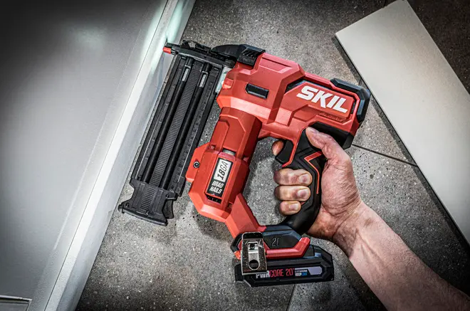 Rød SKIL ledningsfri 16-gauge dykkertpistol med batteri, sort greb og magasin med søm, holdt i hånden på et flisegulv.