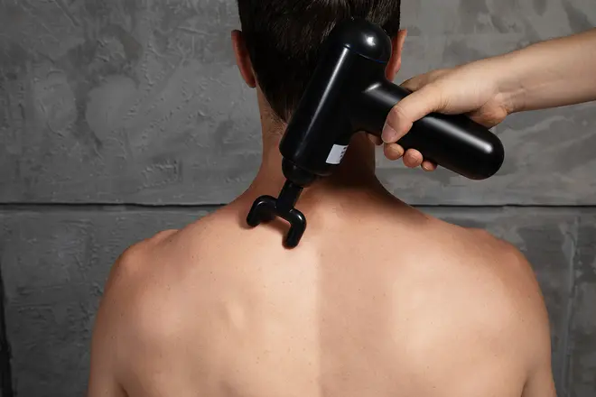 Sort håndholdt massagepistol med gaffeltilbehør bruges på en persons øvre ryg og nakke, designet til muskelafslapning og dybdegående vævsmassage.