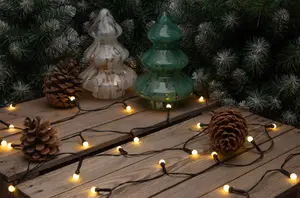 The image shows a festive arrangement featuring two decorative tree-shaped ornaments, one in a glossy white finish with gold accents and the other in a shiny green finish. They are placed on a wooden surface surrounded by pinecones and a string of warm yellow fairy lights. In the background, there are artificial pine branches with a frosted appearance, enhancing the holiday atmosphere.