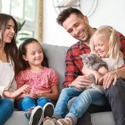 Grå British Shorthair-katt med mjuk päls, runt ansikte och bärnstensfärgade ögon, hålls av ett barn i soffa. Katten ser lugn och frisk ut, perfekt familjedjur.