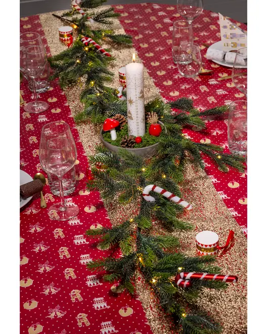 Julebordløber med rødt og guldmønster, pyntet med kunstige grangrene, slikstokke, lyskæder og et centralt stearinlys. Bordet er dækket med glas og bordbomber.