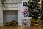 Snögubbe av metallram med LED-lampor, svart hatt, röd fluga och knappar. Står bredvid en eldstad och julgran med inslagna paket och ved.