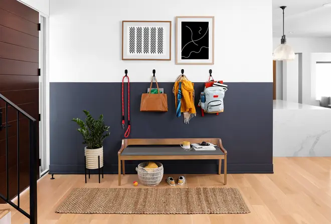 Træbænk med sort sæde, flettet kurv under, kroge over med tasker, jakker og rygsæk. Dekorativ plante, indrammet kunst og flettet tæppe fuldender det moderne udtryk.