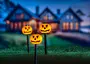 Set med tre Halloween-stigbelysningar formade som leende pumpor på svarta stolpar, lämpliga för utomhusdekoration, med synliga sladdar, lyser orange i mörkret.