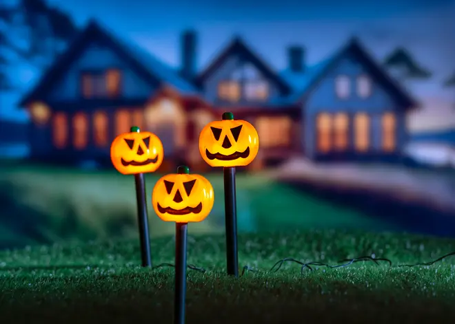 Set med tre Halloween-stigbelysningar formade som leende pumpor på svarta stolpar, lämpliga för utomhusdekoration, med synliga sladdar, lyser orange i mörkret.