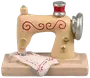 Miniature symaskinefigur, beige med røde og gyldne detaljer, sølvfarvede elementer og et hvidt stofstykke med røde sting, monteret på rektangulær base. Dekorativ samlergenstand.