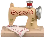 Miniature symaskinefigur, beige med røde og gyldne detaljer, sølvfarvede elementer og et hvidt stofstykke med røde sting, monteret på rektangulær base. Dekorativ samlergenstand.