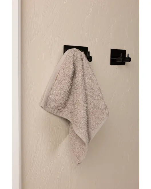 Lys grå håndklæde hænger på en moderne sort vægkrog, monteret på en beige væg med tekstur. Håndklædet ser blødt og absorberende ud, ideelt til badeværelse eller køkken.