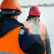 Två arbetare med orange säkerhetsvästar och hjälmar, klädda för kallt väder, står utomhus på en snötäckt arbetsplats. Ånga eller andedräkt syns i luften.