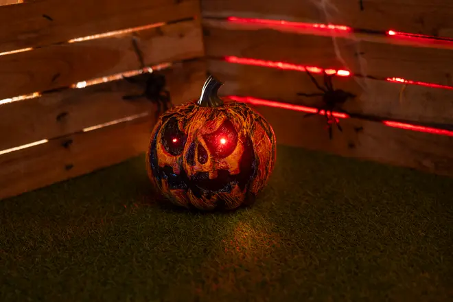 Halloweenpumpa med lysande röda LED-ögon, snidat skrämmande ansikte, texturerad orange yta och svart stjälk. Visas på konstgräs med träbakgrund.