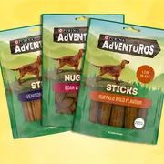Tre Purina Adventuros hundegodbidposer med vildtkødspinde, vildsvinenuggets og bøffelsticks, lavt fedtindhold, uden tilsatte farver og genanvendelig emballage.