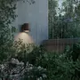 Udendørs væglampe med cylindrisk, mørkebrun finish, monteret på væg, oplyser haveområde med grønne planter og lyserøde blomster, vist i regnvejr.