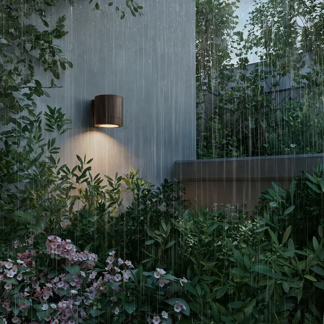Udendørs væglampe med cylindrisk, mørkebrun finish, monteret på væg, oplyser haveområde med grønne planter og lyserøde blomster, vist i regnvejr.