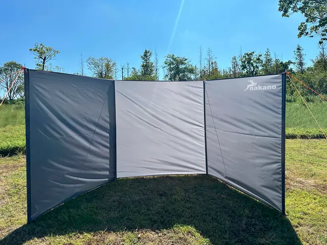 Tredelt udendørs vindskærm med gråt stof og sort kant, fastgjort med snore og pløkker. Giver læ og privatliv til camping eller picnic. Nakano-logo øverst til højre.