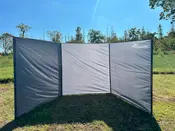 Tredelt udendørs vindskærm med gråt stof og sort kant, fastgjort med snore og pløkker. Giver læ og privatliv til camping eller picnic. Nakano-logo øverst til højre.