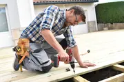 Mand knæler på træterrasse og bruger en ledningsfri boremaskine til at fastgøre brædder. Har værktøjsbælte med hammer, målebånd og kniv. Iført ternet skjorte og arbejdsbukser.
