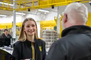 Sort lynlåsjakke med gul kant og gult logo på venstre bryst, båret af en person i et detail- eller lagerområde.