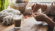 Person strikker med cremefarvet garn, omgivet af garnnøgler og kurv på et træbord med et glas kaffe. Hyggelig, hjemlig atmosfære til håndarbejde.