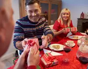 Röd och vitrandig presentask med rött band, överlämnas vid ett festligt dukat bord med vinglas, tallrikar och juldekorationer.