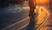 En person cykler langs sporvognsskinner på en brostensbelagt gade ved solnedgang, hvor det varme sollys reflekteres i skinner og belægning.