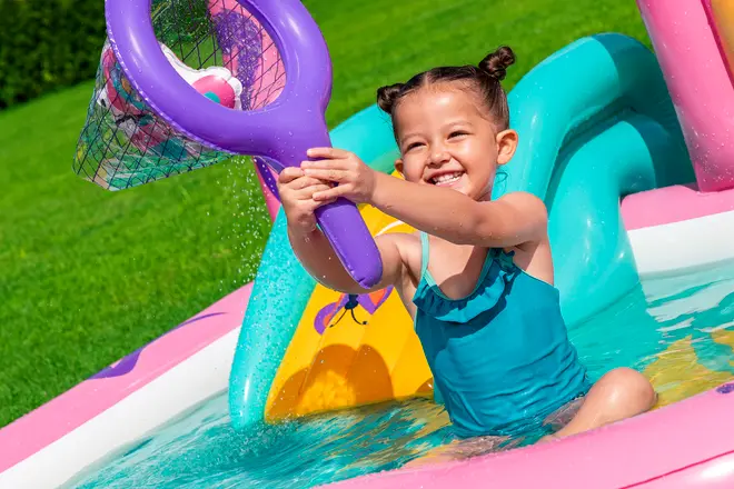 Barn i blå badedragt leger i oppustelig pool med stor lilla net og fanger legetøj. Poolen har farverige rutsjebaner og vandaktiviteter, ideel til udendørs sommerleg for små børn.