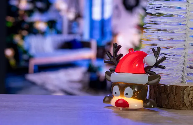 Lille rensdyrfigur med rød julemandskasket, brune gevirer og store øjne. Figuren er oplyst indefra og står ved siden af et hvidt kunstigt juletræ på træoverflade.