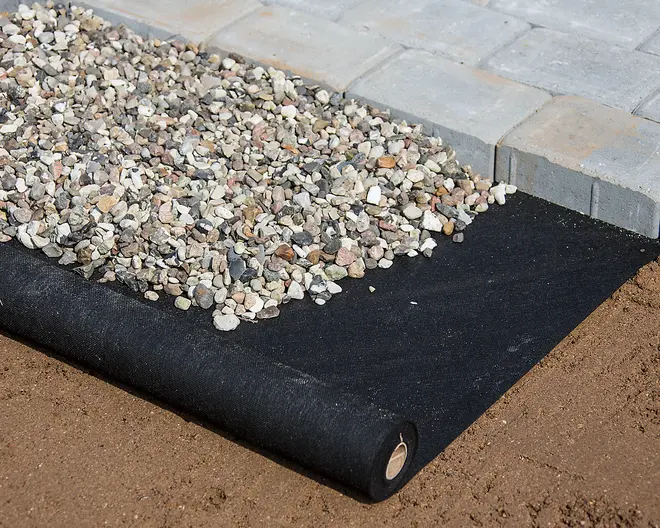 Svart markduk utrullad på jord, delvis täckt med grus, intill betongplattor. Används för ogräskontroll och dränering i trädgårdsprojekt.