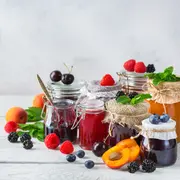 Assorterede glas med hjemmelavet frugtmarmelade, pyntet med friske bær eller urter, omgivet af hele frugter som abrikoser, kirsebær, hindbær og brombær på hvidt træbord.