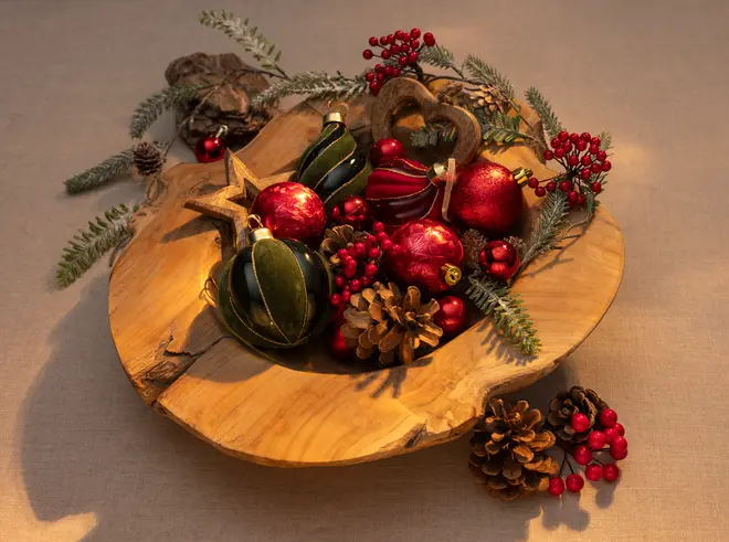Træskål fyldt med julepynt, kogler, røde bær, grønne og røde kugler, guldfarvet stjerne og grangrene. Festlig dekoration til bordet.
