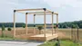 Trærammekonstruktion med metalbeslag, hævet platformbund, åbne sider og top, velegnet til opbygning af et lille udendørsrum eller pavillon. Placeret på græs med flisebelægning.