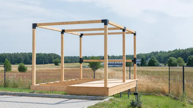 Trærammekonstruktion med metalbeslag, hævet platformbund, åbne sider og top, velegnet til opbygning af et lille udendørsrum eller pavillon. Placeret på græs med flisebelægning.