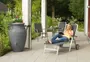 Stor grå regntunna för utomhusbruk med handtag och blomplantering upptill. Placerad på uteplats bredvid utemöbler, dekorativ och funktionell för regnvatteninsamling.