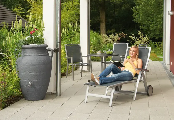 Stor grå regntunna för utomhusbruk med handtag och blomplantering upptill. Placerad på uteplats bredvid utemöbler, dekorativ och funktionell för regnvatteninsamling.