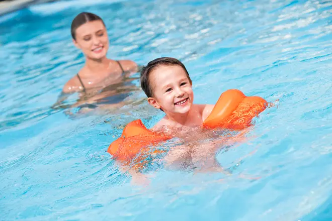 Barn med orange oppustelige svømmevinger i en swimmingpool. Svømmevingerne giver opdrift og sikkerhed, ideelle til børn, der lærer at svømme. Lavet af slidstærkt, vandtæt materiale.