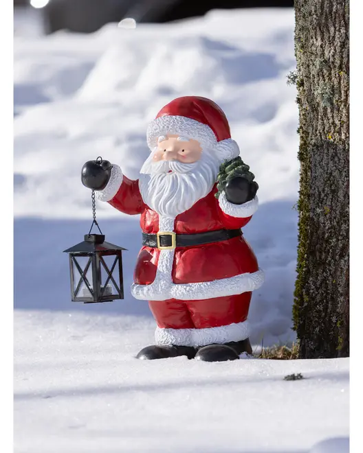 Festiv julemandfigur i rødt outfit med sort lanterne, til udendørs juledekoration, stående i sne ved et træ.
