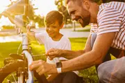 Voksen og barn reparerer en hvid cykel udendørs med en skruetrækker ved forhjulet. Voksen har stribet trøje og smartwatch. Grøn græs og sollys i baggrunden.